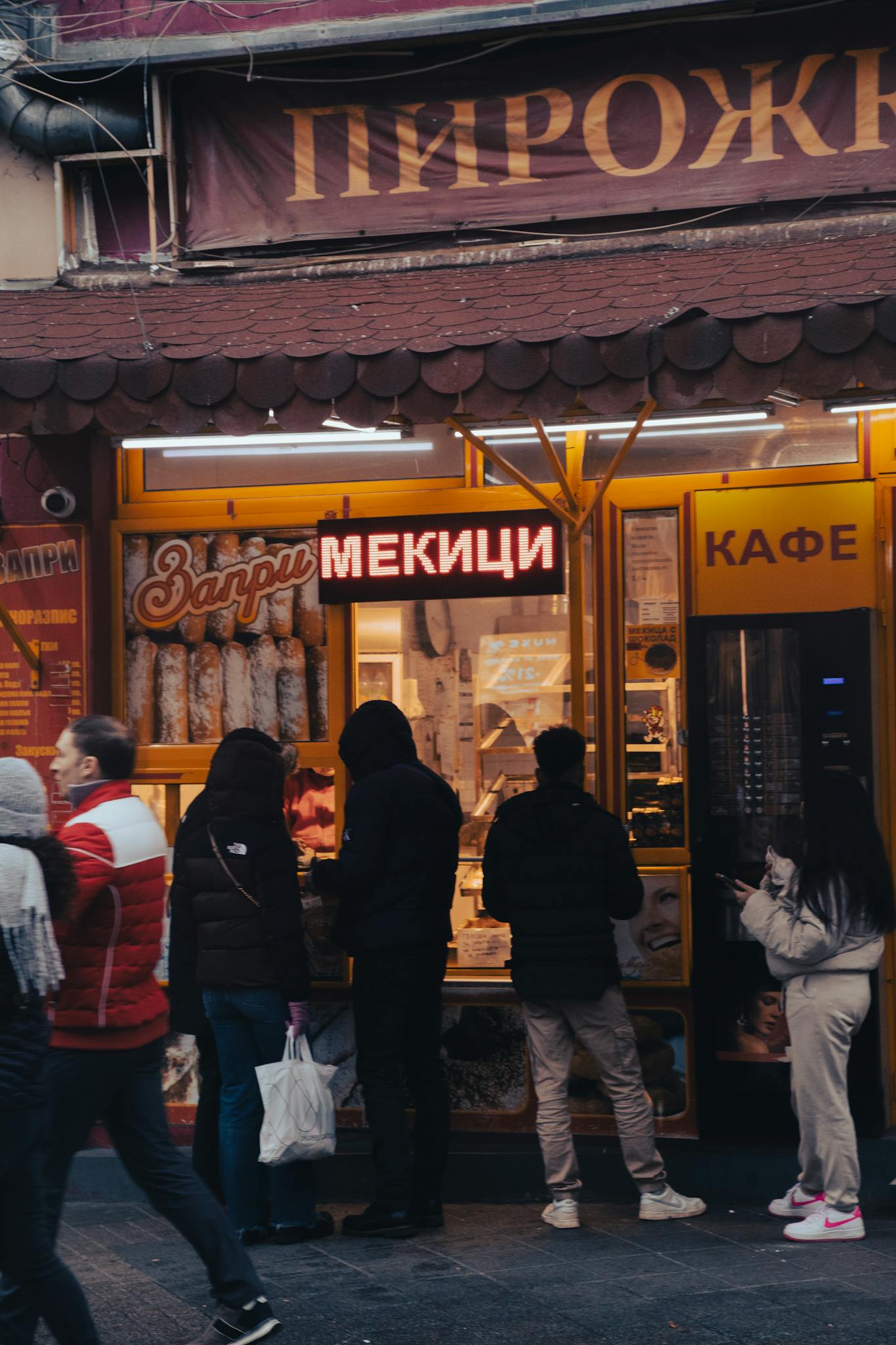 People queue outside a bakery in an Eastern European city, featuring neon signs.